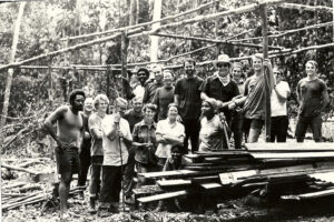 Black-and-white photo of a multiracial group of men, women, and children standing at a construction site in the Guyanese jungle, early Jonestown settlers.