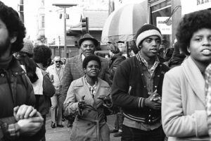 Black-and-white street scene showing diverse protesters marching and singing during the International Hotel eviction protests in San Francisco.
